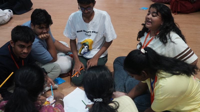 A group on the floor bends over chart paper with pencils while one student makes her point.