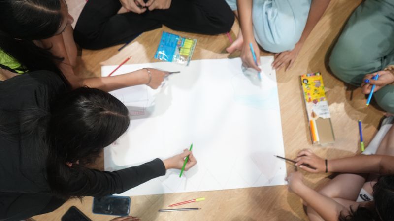 Seen from above, students’ hands drawing with coloured pencils on one large sheet of chart paper.