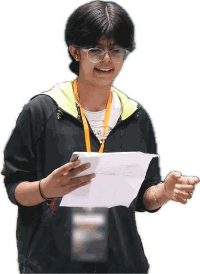 A student in a black hoodie laughs as she reads from the paper in her hands.