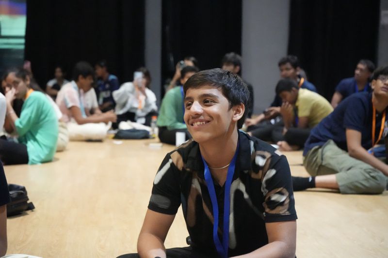 A student sitting on the floor of a busy hall smiles at someone across the circle.