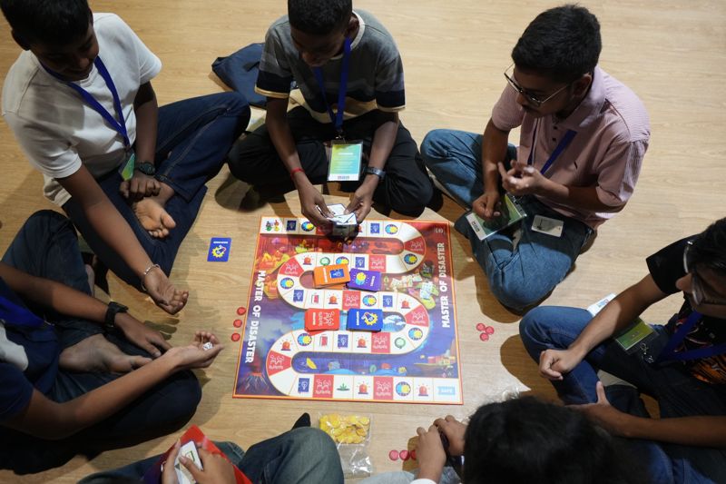 Seen from above, students sit in a circle on the floor around a board game called Master of Disaster, cards in hand.
