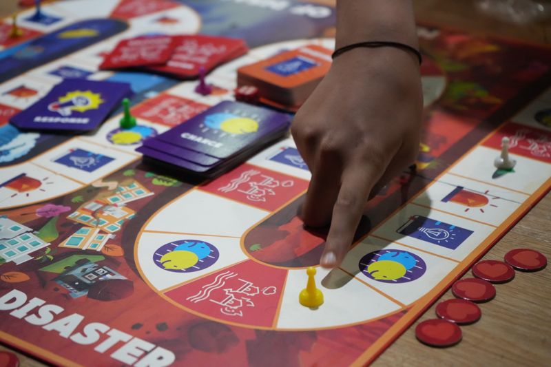 A student’s finger moves a yellow piece along a board game called Disaster, with Response and Chance cards on the board.