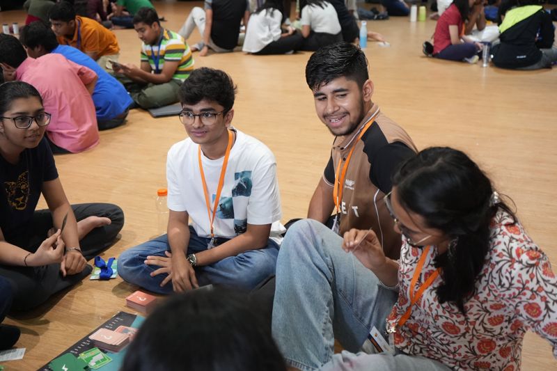 Students sitting cross-legged on the floor smile and talk in a small group.